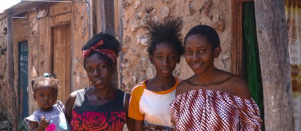 An image of Nalucha holding her daughter, a little girl, in her arms. She flanked by her sisters, in front of her home